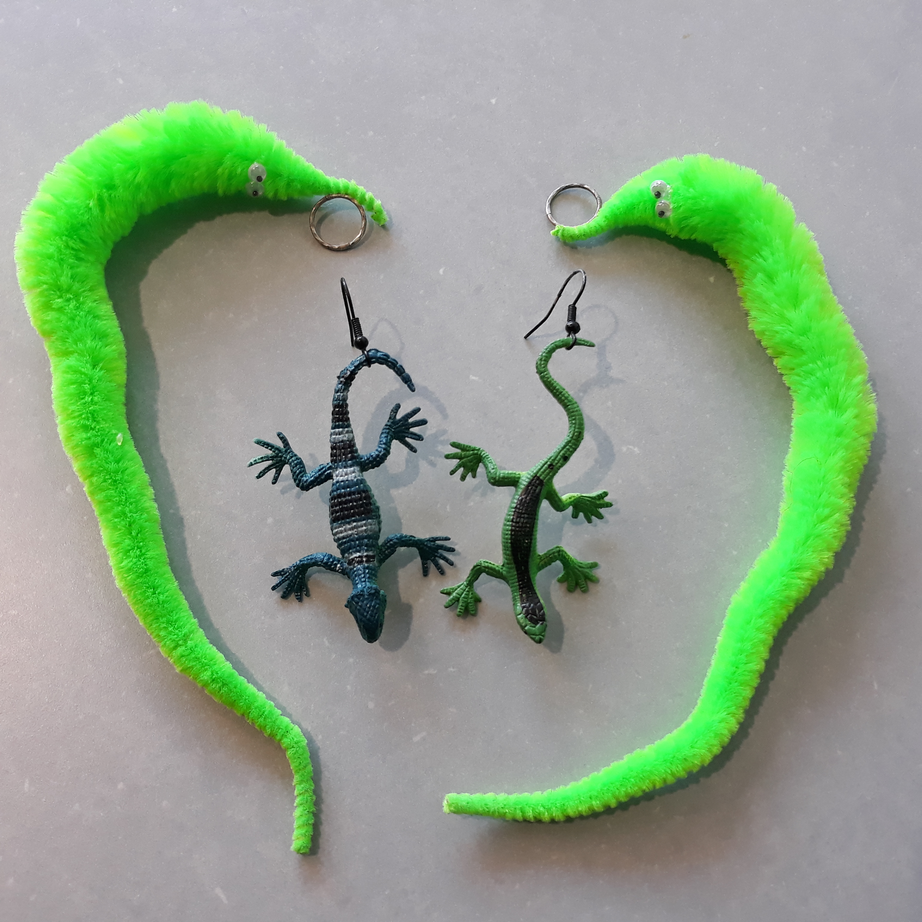 In the middle are two toy lizard earrings, one blue-green with black and white stripes. The other is green with a black stripe running down its back. Around them forming an almost heart shape are bright green worm-on-a-string earrings.