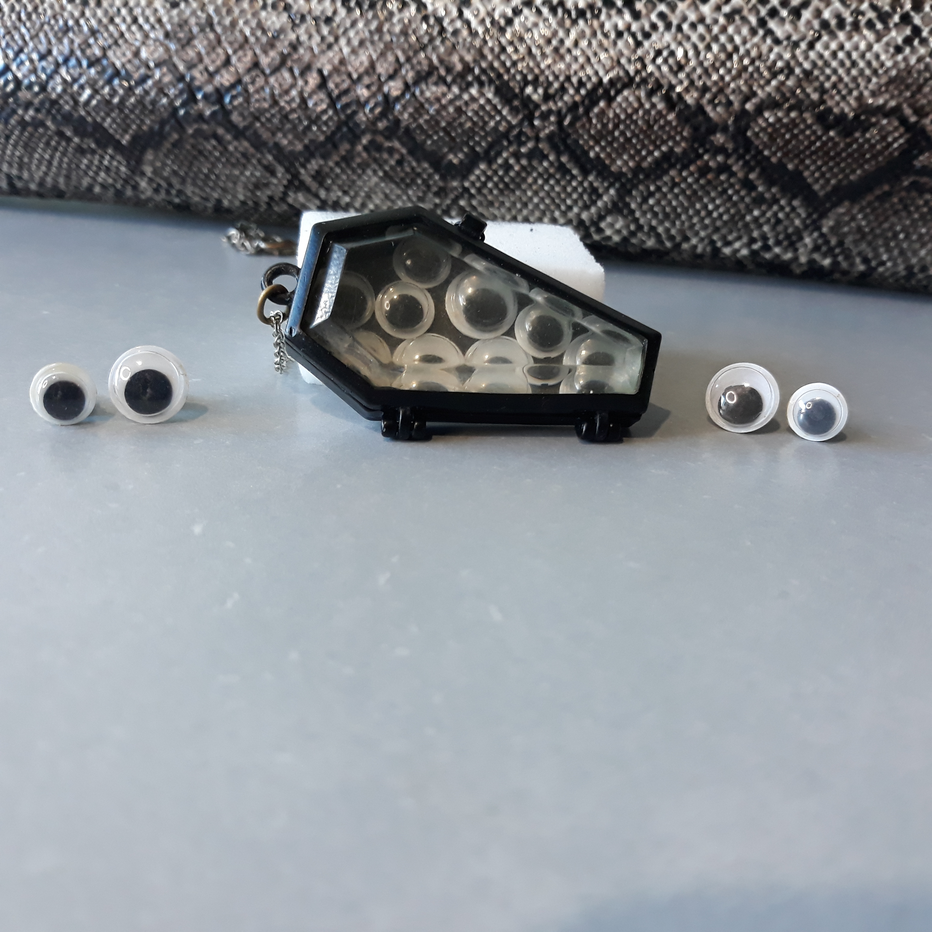 A necklace is in the middle, with the pendant as the focus. It is a black coffin with a glass lid. Inside are a bunch of googly eyes. On either side are two sets of earrings, one big googly eye each and one small.