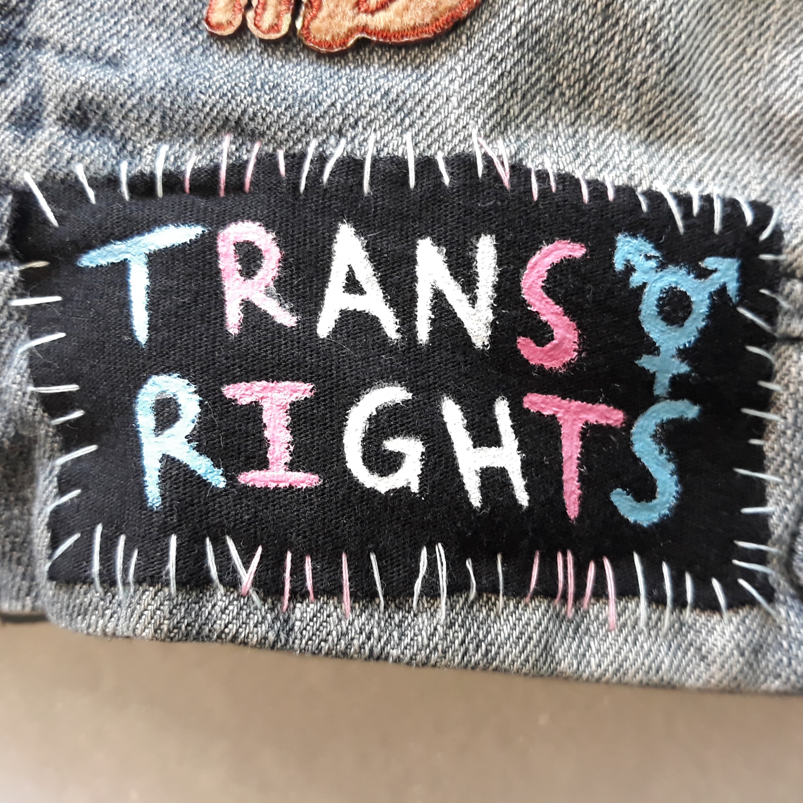 A patch that has 'trans rights’ with the trans symbol drawn next to 'trans’. The writing and thread changes between blue, pink and white to mimic the trans flag.