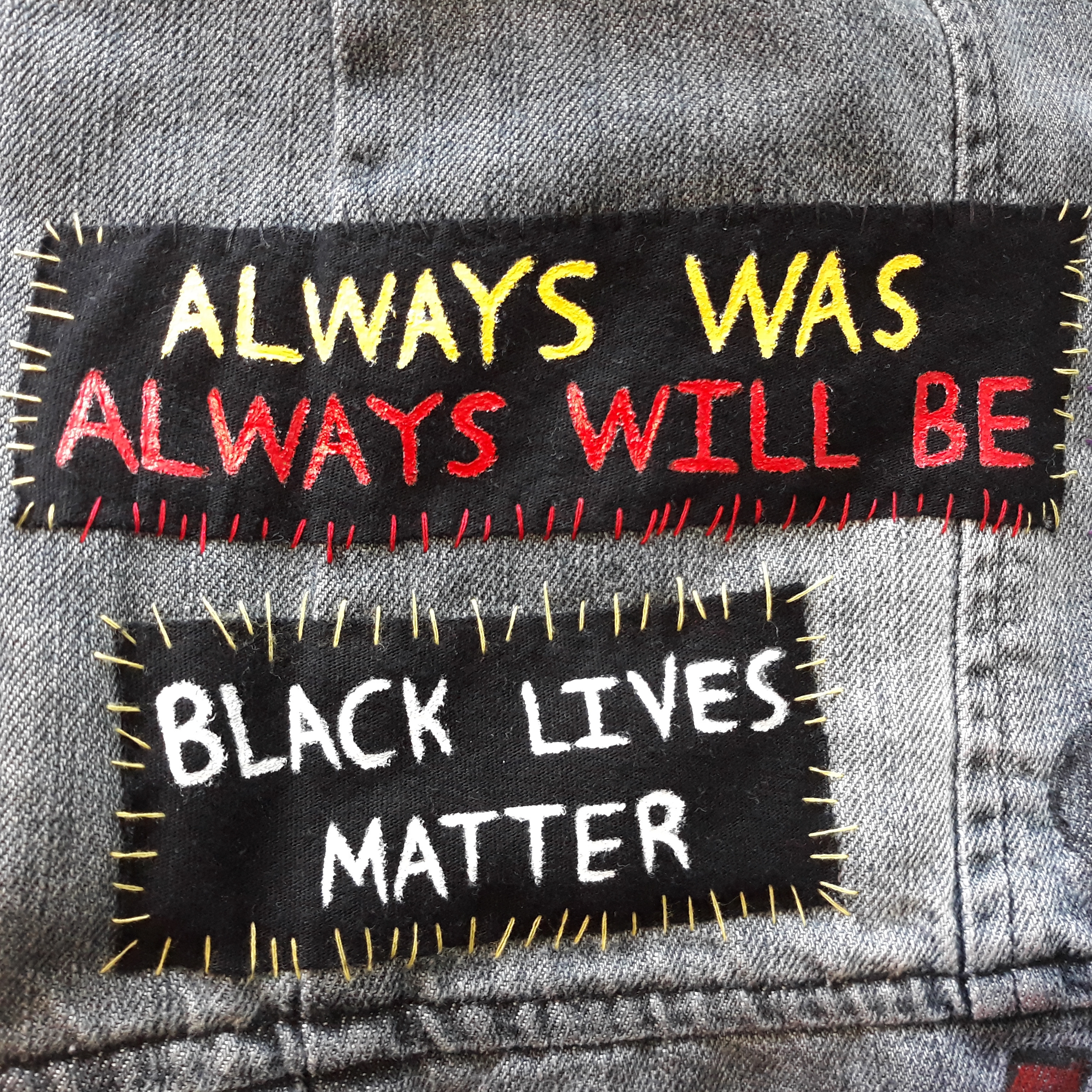 Two hand painted patches. The top one reads ‘always was’ in yellow, above 'always will be’ in red. It is sewn on with red, black and yellow thread. The bottom one reads 'Black lives matter’. It is written in white and sewn on with yellow thread.