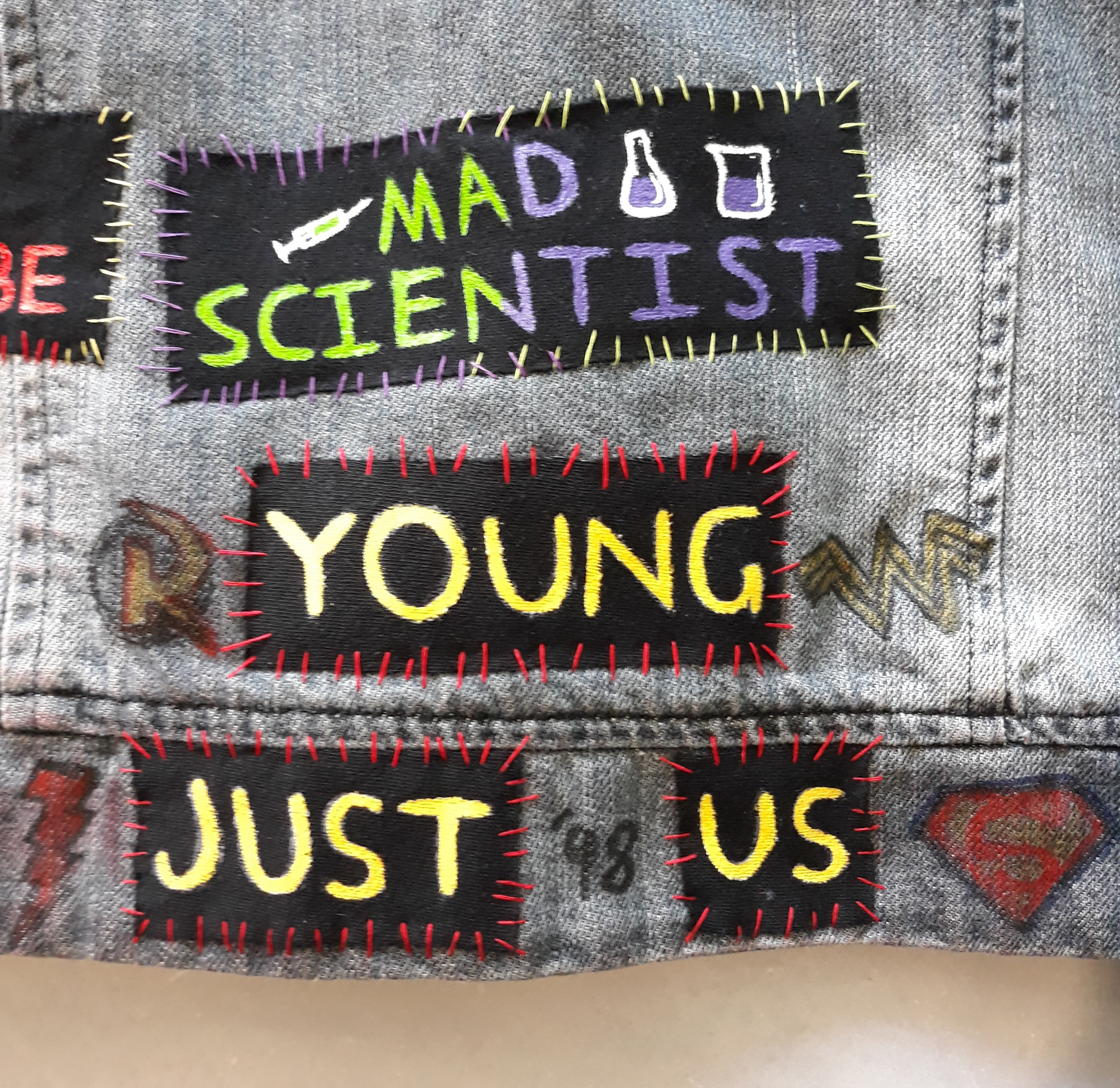 Multiple hand painted patches. The top patch reads ‘mad scientist’, with one half green and the other half purple. Next to 'mad’ is a syringe filled with green liquid on the left. On the right filled with purple liquid is a conical flask and beaker. It is sewn on with green and purple thread. Below that are three patches separating out the words 'young just us’. The writing is yellow and they are sewn on with red thread. Surrounding these are various symbols drawn on the denim with fabric pen. These are Robin’s symbol, Wonder Girl’s symbol, a red lightning bolt for Impulse, the words '98 and an S-shield for Superboy.