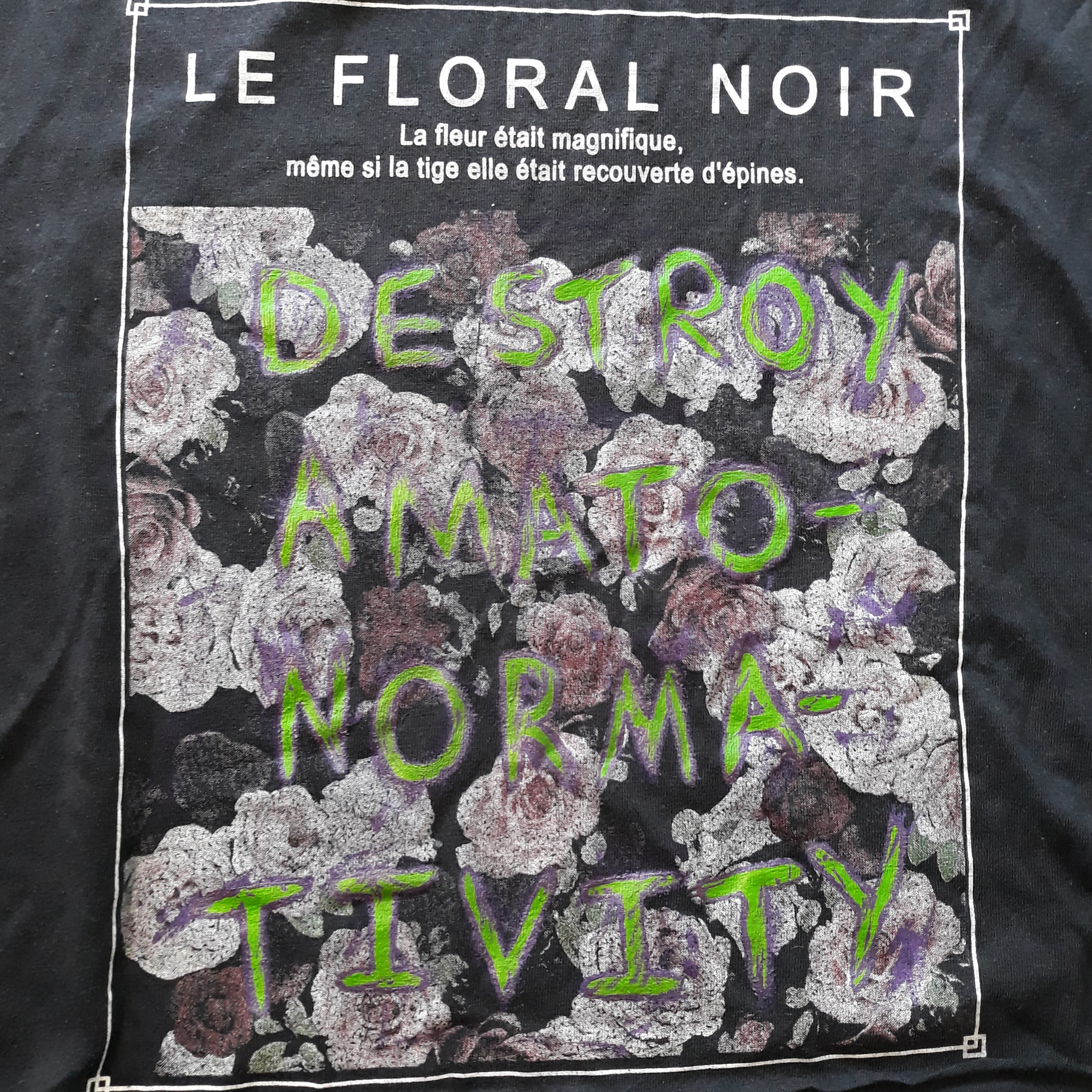 Close up photo of a black shirt. Printed in the middle are realistic white, pink and purple roses, with writing in French above them. Hand-painted over the flowers is ‘DESTORY AMATO-NORMA-TIVITY’. It is written in bright green with a messy purple outline.